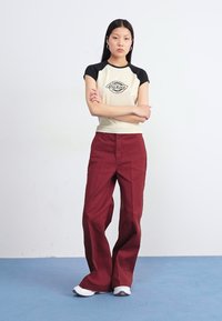 Junge Frau steht mit verschränkten Armen da und trägt ein cremefarbenes Shirt mit schwarzen Ärmeln und weiten, kastanienbraunen Hosen in einem minimalistischen Studio.