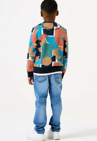 Bambino in piedi di spalle, che indossa un maglione colorato con un motivo geometrico, jeans blu e sneakers bianche su uno sfondo semplice.