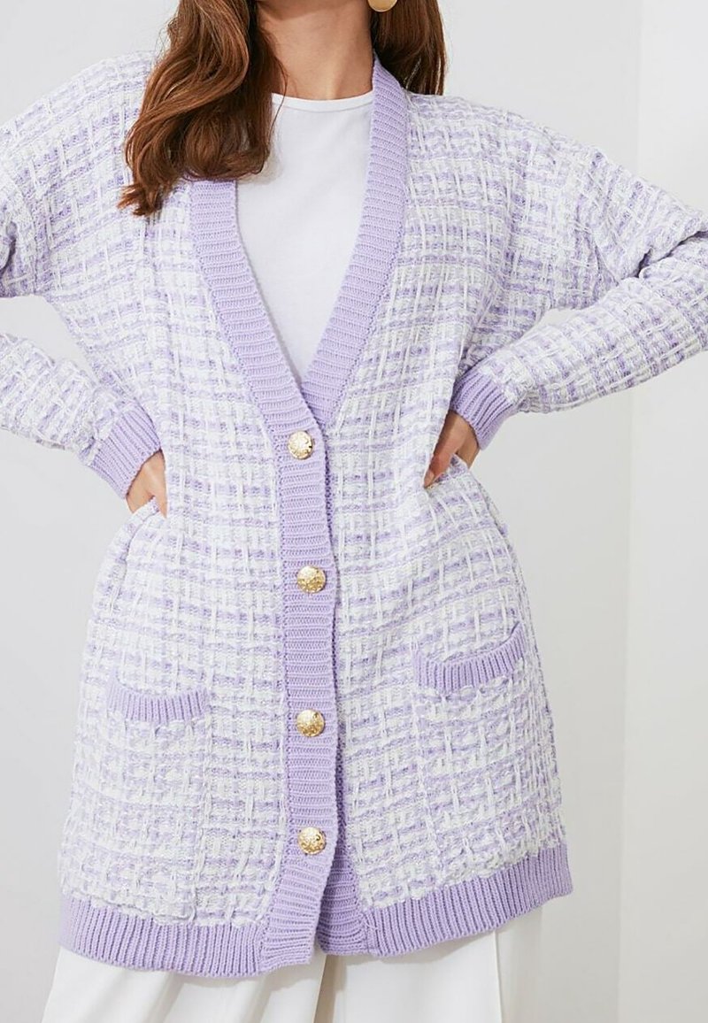 Femme portant un long cardigan texturé lavande et blanc avec des boutons dorés, des finitions côtelées et des poches avant sur un haut blanc.