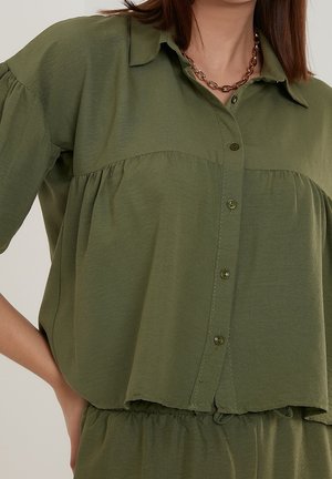 Vrouw draagt een loszittend olijfgroen overhemd met knopen en geplooide naden, en een dikke kettingketting, hand rust op de heup.