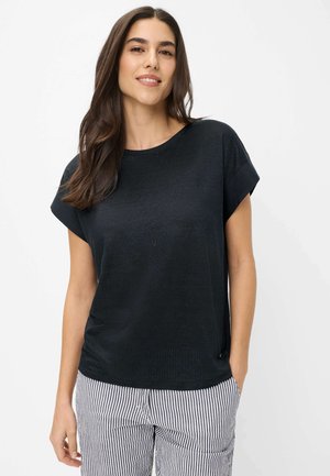 Kvinde med langt mørkt hår iført en sort top med korte ærmer og hvide bukser med sorte lodrette striber, stående foran en hvid baggrund.