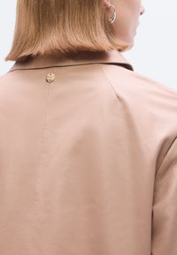 Šviesiai smėlio spalvos odinė striukė, pasižyminti lygiu paviršiumi, klasikine apykakle ir auksinės spalvos apvalia detalė nugaroje ties kaklu.