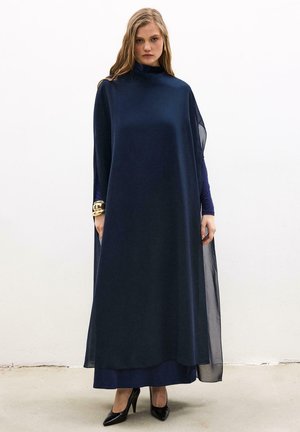 Femme aux longs cheveux blonds portant une robe cape bleu marine jusqu'au sol et des talons noirs, debout contre un mur blanc uni.