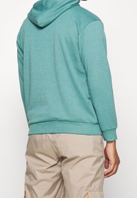 Felpa turchese con una texture morbida, tasca a marsupio e cappuccio regolabile con cordino. Indossata con pantaloni cargo beige con accenti arancioni.