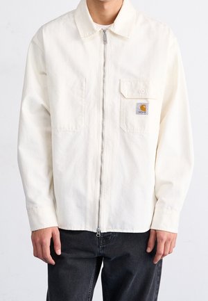 Man draagt een wit jack met ritssluiting en een borstzak met Carhartt-logo, gecombineerd met een zwarte broek, tegen een effen achtergrond.