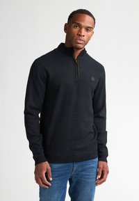 Maglione nero a maglia con colletto alto e zip frontale a metà. Presenta un motivo testurizzato e un piccolo logo sul petto. Abbinato a jeans blu.