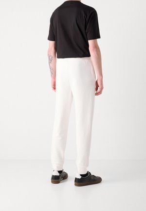 Osoba ubrana w czarny t-shirt, białe spodnie, czarne trampki z białymi piętami, stojąca tyłem do kamery na białym tle.