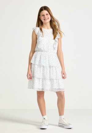 Smilende pige med langt hår iført en hvid ærmeløs kjole med blomstret print og flæser, kombineret med hvide sneakers og sokker.