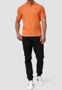 T-shirt polo orange avec un col et deux boutons, associé à un pantalon cargo noir et des baskets blanches avec des accents noirs.