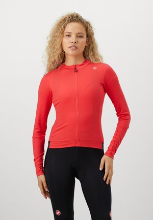Frau mit lockigem blonden Haar, die ein rotes langärmliges Radtrikot mit Reißverschluss und schwarze gepolsterte Radsportshorts trägt.