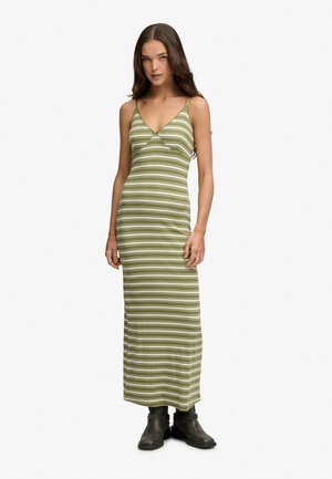 Kobieta z długimi brązowymi włosami, ubrana w zielono-białą, paskowaną, bezrękawnikową sukienkę maxi i czarne botki, stojąca prosto.