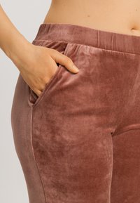 Měkké sametové kalhoty v růžovo-hnědé barvě s elastickým pasem a bočními kapsami, které zdůrazňují hladký povrch a dobře padnoucí střih.