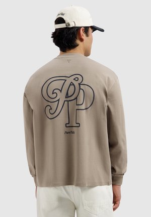 Man wearing beige long-sleeve shirt with large "PP" initials and "Pure Path" text on the back, white pants, and white and black cap.