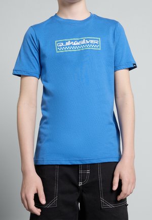 Dziecko nosi niebieską koszulkę Quiksilver i czarne spodenki z białymi przeszyciami, stoi na tle jednolitego szarego tła.