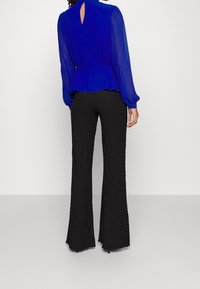 Blue, long-sleeve blouse with a keyhole cutout at the back, worn with black textured flared pants. Smooth fabric contrasts with rich texture.