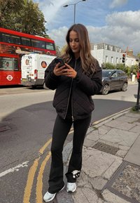 Fekete steppelt dzseki cipzárral, fekete bővülő nadrág grafikai mintával és ezüst színű tornacipők. Városi környezet, háttérben piros busz látható.