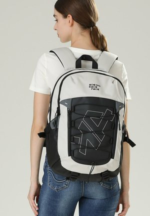 Femme portant un sac à dos blanc et noir au design géométrique, un t-shirt blanc et un jean bleu, tournée de dos sur un fond uni.