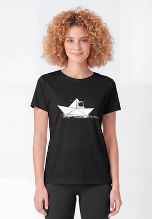 Jeune femme aux cheveux bouclés portant un t-shirt noir avec un bateau en papier blanc et un astronaute flottant sur l'eau.