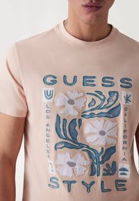 Světle růžové tričko s květinovou výšivkou v bílém a modrém provedení, s textem "GUESS" a čtvercovým grafickým uspořádáním. Krátké rukávy.