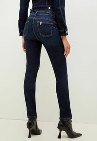 Tunna mörkblå jeans med bakfickor, kontraststygn och hög midja; kombinerat med svarta blanka klackstövlar.