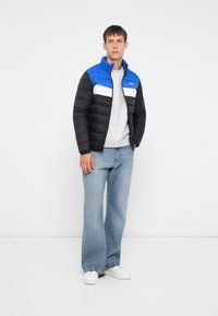 Polstret jakke i sort og blå med en hvid stribe hen over brystet, lynlås lukning og høj krave. Sammen med lyseblå jeans og sneakers.