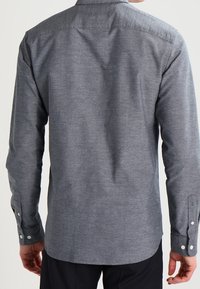 Šedá tkaná košile s dlouhými rukávy, manžetami na knoflíky a límcem. Má jemnou texturu a zakřivený lem na zadní straně.