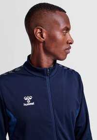 Marinblå zip-up sportjacka med texturerad yta, med ljusblå accenter och en logga på bröstet. Svart mönstrad detalj på axlarna.