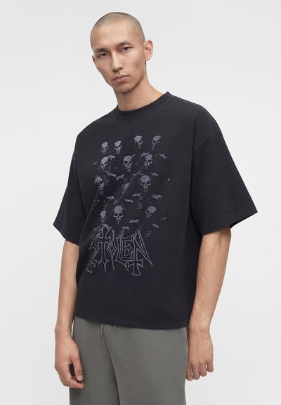Mann mit rasiertem Kopf trägt ein schwarzes oversized T-Shirt mit mehreren Skelett-Grafiken und stilisiertem Text, kombiniert mit grauer Hose.