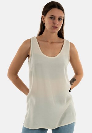Mujer con largo cabello castaño, vestida con un top blanco sin mangas y vaqueros azules, de pie con las manos detrás de la espalda frente a un fondo liso.