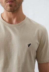 Camiseta de color beige claro hecha de tejido suave, con cuello redondo y un pequeño bordado de un pájaro negro en el pecho.