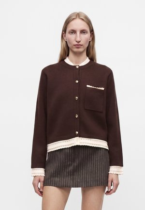 Femme aux cheveux blonds et raides porte un cardigan marron avec des boutons dorés et une jupe à fines rayures, debout devant un fond clair uni.