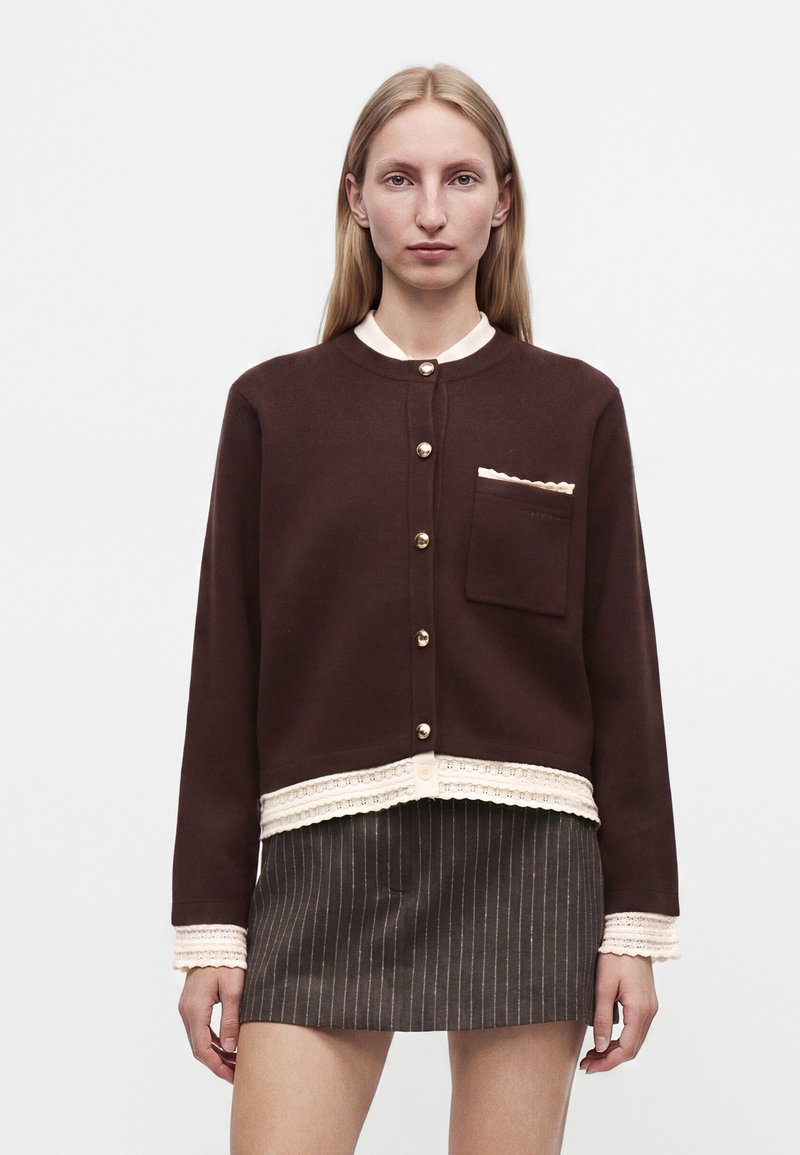 Femme aux cheveux blonds et raides porte un cardigan marron avec des boutons dorés et une jupe à fines rayures, debout devant un fond clair uni.