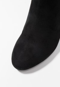 Bottine en daim noir avec un bout pointu et une texture lisse. La partie supérieure présente un design épuré sans éléments métalliques visibles.