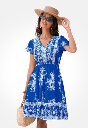 Kobieta w niebieskiej sukience w kwiaty, szerokim słomkowym kapeluszu, okularach przeciwsłonecznych i trzymająca plecioną torebkę, pozująca na jednolitym tle.