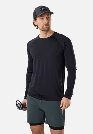 Mand iført sort langærmet skjorte, grønne shorts med sort kompressionslag, sort kasket, holder en sort vandflaske og kigger mod venstre.