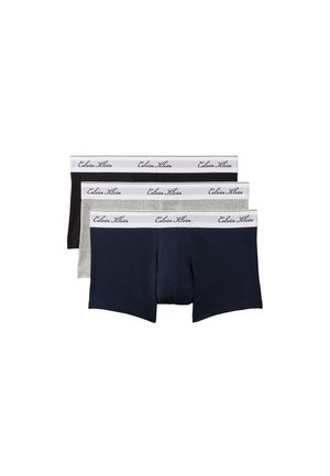 Młody mężczyzna stoi boso, ubrany w szare bokserki Calvin Klein na jednolitym szarym tle.