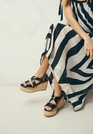 Frau sitzt mit übereinandergeschlagenen Beinen, trägt ein schwarz-weiß gestreiftes Kleid und schwarze Keilsandaletten mit Riemchen um den Knöchel.