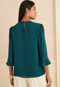 Modrá blúzka s plisovanými rukávmi a skladanými manžetami. Detail s kľúčovou dierou na zadnej strane, mäkká tkanina a čipkované akcenty na okrajoch rukávov.