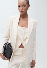 Slonovinový šitý blazer přes odpovídající zapínací top a přiléhavé kalhoty, s černou kabelkou v ruce. Hladká tkanina, klasický design, minimální akcenty.