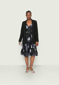 Blazer preto sobre um vestido floral escuro com tons de cinza e roxo, com comprimento até ao joelho e um corpete ajustado, acompanhado de saltos prateados e uma clutch.