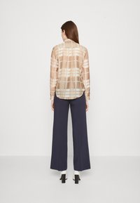 Gennemsigtig, langærmet beige bluse med et rutenetmønster i lysere nuancer, parret med marineblå vide bukser og hvide blokhælede sko.
