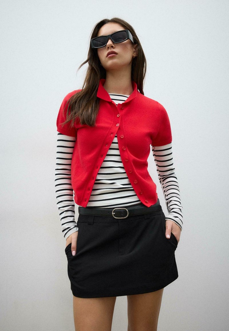 Femme portant des lunettes de soleil noires, un cardigan rouge à manches courtes par-dessus un haut à manches longues rayé noir et blanc, jupe noire avec ceinture, mains dans les poches.