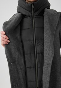 Culoare gri, pardesiu din lână, evidențiind o geacă puffer neagră cu guler înalt. Are un fermoar frontal și detalii fine de cusătură.