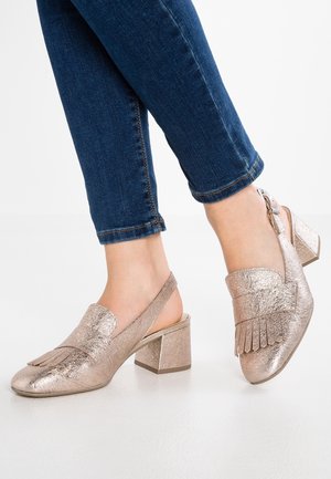 Metaliczne różowe złote buty na obcasie z luźnym przodem, frędzlami i masywnym słupkowym obcasem. Noszone z ciemnoniebieskimi dżinsami.