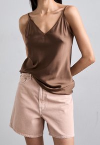 Camiseta de satén marrón con escote en V y tirantes finos, combinada con pantalones cortos de mezclilla rosa claro. La tela parece suave y lisa.