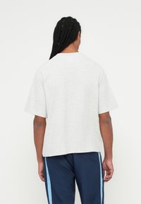 Uomo con lunghi capelli intrecciati che indossa una semplice maglietta grigia chiara a maniche corte e pantaloni blu navy con strisce azzurre, di spalle.