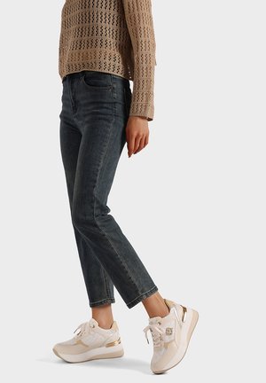 Sneakers con una combinazione di colori beige e bianco, materiali lisci e testurizzati, e accenti metallici; abbinate a jeans aderenti di colore blu scuro.