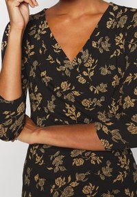 Black wrap dress featuring a floral pattern in gold, with three-quarter sleeves and a deep V-neckline. Soft, textured fabric.