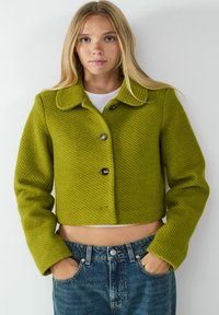 Veste courte vert lime en tissu texturé avec col arrondi et quatre boutons à l'avant ; portée sur un haut blanc et un jean bleu.
