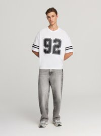 Biały krótki rękaw koszulki z czarnymi paskami i numerem 92 z przodu; zestawiony z jasnoszarymi prostymi dżinsami i szarymi tenisówkami.
